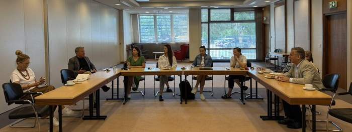 Pokaż zdjęcie: Kilka osób siedzi przy stołach ustawionych w kształt litery U w jasnej sali konferencyjnej. W tle duże okna z widokiem na drzewa i czerwone fotele. Na stołach napoje, filiżanki i dokumenty.