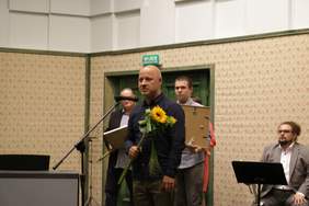 Pokaż zdjęcie: Przed mikrofonem stoi łysy mężczyzna, ubrany w granatową koszulę w białe kropki. Trzyma bukiet kwiatów. Z tyłu stoi dwóch mężczyzn obaj trzymają dyplomu. Na lewo od przemawiającego siedzi trębacz, ubrany w jasny garnitur.