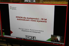 Pokaż zdjęcie: Kadr na monitor z trwającego połączenia online z Międzynarodowej Konferencji Państwowego Funduszu Osób Niepełnosprawnych. Uśmiechnięta kobieta w okularach, w prawym, dolnym rogu ekranu. 
