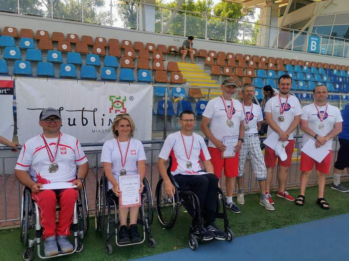 Pokaż zdjęcie: Na wózkach siedzą dwaj mężczyźni i jedna kobieta z medalami na szyi. Kobieta trzyma w ręku dyplom. Dalej w rzędzie stoją czterej mężczyźni z medalami na szyi. W tle stadion