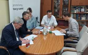 Pokaż zdjęcie: siedziba Oddziału w tle duży baner z logo PFRON. Przy owalnym stole siedzi czterech mężczyzn i podpisuje dokumenty i umowę o dofinansowanie ze środków PFRON