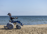 Osoba siedzi na specjalistycznym wózku plażowym z dużymi, szerokimi kołami na piaszczystej plaży. W tle widoczne jest morze i jasne niebo. Wózek ustawiony jest bokiem.