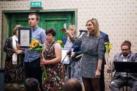 Pokaż zdjęcie: Na pierwszym planie trzy osoby, mężczyzna z dyplomem w ręku, kobieta w sukience ze słonecznikiem, pani kierownik ZAZ w biało-czarnej sukience ze statuetką w dłoni. W tle inni laureaci i orkiestra