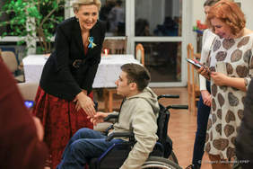 Pokaż zdjęcie: Na zdjęciu są 4 osoby, kobieta pani prezydentowa pochyla się uśmiechnięta i wyciąga do rękę do chłopca siedzącego na wózku inwalidzkim. Za wózkiem po prawej stronie zdjęcia stoją dwie uśmiechnięte kobiety.