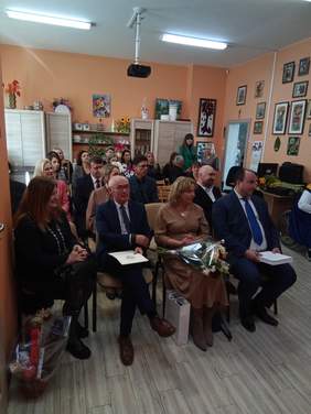 Pokaż zdjęcie: W sali domu opieki na krzesłach siedzą zaproszeni goście, w pierwszym rzędzie mężczyźni w garniturach oraz kobieta w beżowej sukience trzymająca kwiaty