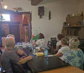 Pokaż zdjęcie: Grupa seniorek uczestniczy w spotkaniu informacyjno-konsultacyjnym w Klubie Seniora 60+ w Zaściankach. Uczestniczki siedzą przy stole i słuchają prezentacji na temat programów PFRON. Na ścianie widoczny jest plakat informacyjny Funduszu, a na stole znajdują się dokumenty oraz ulotki.