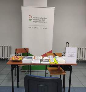 Pokaż zdjęcie: Zdjęcie przedstawia puste stoisko wystawiennicze Oddziału Zachodniopomorskiego Państwowego Funduszu Rehabilitacji Osób Niepełnosprawnych (PFRON). Na pierwszym planie widać ciemne krzesło biurowe tyłem do obiektywu. Za nim stoi prostokątny stolik, na którym równo poukładano materiały informacyjne, ulotki, smycze oraz miskę z kolorowymi cukierkami. Po prawej stronie blatu stoi biała tabliczka z nazwą instytucji. Za stolikiem znajduje się duży biały roll-up z logo PFRON (stylizowany kwiat tulipana) i adresem strony internetowej. Tło stanowią jasne ściany i białe grzejniki żebrowe.