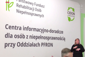 Pokaż zdjęcie: Zbliżenie na białą ściankę z logotypami i napisami w języku polskim. Widoczne logo PFRON (zielony kwiatek) oraz logo CIDON (Centra informacyjno-doradcze dla osób z niepełnosprawnością). Duży napis głosi: „Centra informacyjno-doradcze dla osób z niepełnosprawnością przy Oddziałach PFRON”. W prawym dolnym rogu znajduje się tłumaczka języka migowego w czarnej bluzce, dynamicznie gestykulująca.