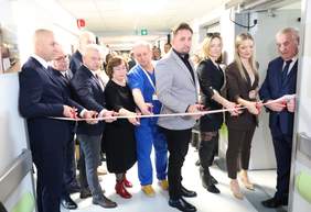 Pokaż zdjęcie: Grupa osób bierze udział w ceremonii przecięcia wstęgi. Na pierwszym planie widoczny jest mężczyzna w garniturze, trzymający nożyczki, otoczony przez innych uczestników w eleganckich ubraniach. Wszyscy trzymają nożyczki i czekają na symboliczne przecięcie wstęgi, otwierając nowe pomieszczenie.