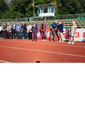 Na stadionie, na bieżni w szeregu stoi kilkanaście osób, mężczyzna w garniturze i ciemnych okularach przemawia