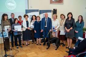 Pokaż zdjęcie: Na scenie grupa dwunastu osób pozuje do pamiątkowego zdjęcia, pośrodku stoi minister Wdówik, a przed nim jest pies przewodnik. 
