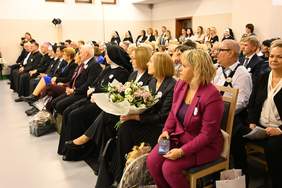 Pokaż zdjęcie: Na krzesełkach siedzą zaproszeni goście, kilka osób trzyma w ręku kwiaty.