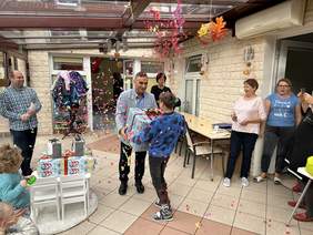 Pokaż zdjęcie: Mężczyzna w ciemnych spodniach jasnej marynarce przekazuje prezent chłopcu w granatowym sweterku. Prezent jest bardzo duży opakowany w kolorowy papier. Do dokoła nich fruwają kolorowe konfetti. Wszyscy są uśmiechnięci. 