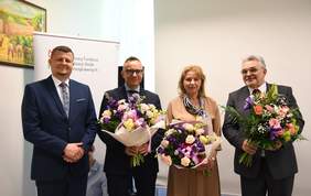 Pokaż zdjęcie: Na zdjęciu cztery osoby, trzy z nich trzymają duże bukiety kolorowych kwiatów. Mężczyźni są ubrani w garnitury, kobieta stojąca jako trzecia ma jasną beżową sukienkę