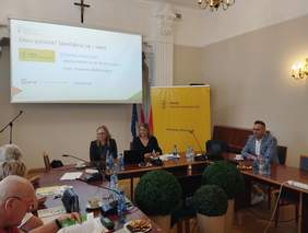 Pokaż zdjęcie: Przy stołach ustawionych w okrąg siedzą ludzie. Nad ich głowami wisi tablica multimedialna, na której wyświetlone są zagadnienia dotyczące dostępności. Na stołach leżą dokumenty, ciastka i filiżanki z kawą. 