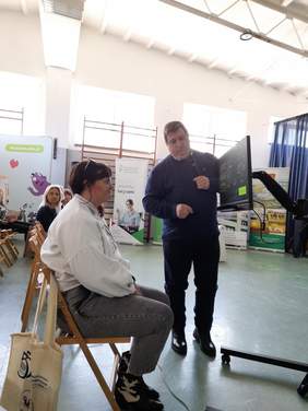 Pokaż zdjęcie: Na krześle przed monitorem siedzi kobieta, która wpatruje się w monitor. Obok niej stoi mężczyzna prelegent, tłumaczy działanie urządzenia PC-Eye.