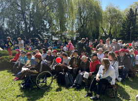 Pokaż zdjęcie: Na polanie siedzą ludzie na ławkach w rzędach. Na pierwszym planie widać chłopca siedzącego na wózku inwalidzkim w tle drzewa wysokie i trzy krzyże.