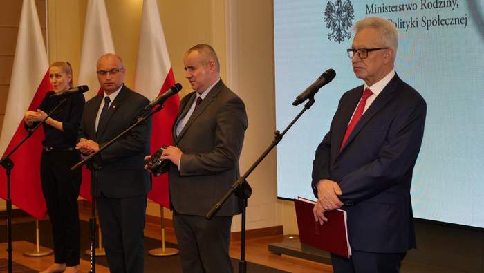 Pokaż zdjęcie: Konferencja prasowa, prelegenci na tle flag biało-czerwonych, ekran z napisem Ministerstwo Rodziny, Pracy i Polityki Społecznej. Tłumaczka jerzyka migowego. 