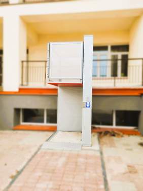 Pokaż zdjęcie: Na tle jasnożółtego budynku jest podnośnik pionowy w szarym kolorze. Jest on na wysokości parteru od balkonu.