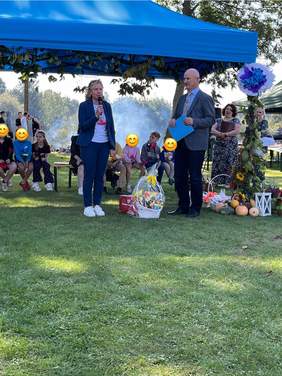 Pokaż zdjęcie: W plenerze, na trawie stoją dwie osoby, kobieta ubrana w garnitur i trampki trzyma w ręku mikrofon, obok stoi mężczyzna w garniturze. Kosz słodyczy stoi na trawie.