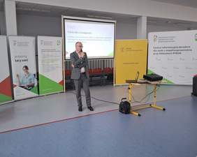 Pokaż zdjęcie: Mężczyzna w garniturze stoi z mikrofonem, za jego plecami na ekranie wyświetlany slajd "Polski Akt o Dostępności". W tle widoczne są banery PFRON. 