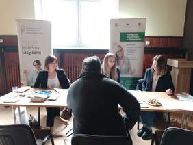 Pokaż zdjęcie: Trzy kobiety siedzą przy stole na którym leżą broszury i informatory przed nimi na krześle siedzi mężczyzna. Za kobietami stoją dwa banery z logo PFRON i PUP.