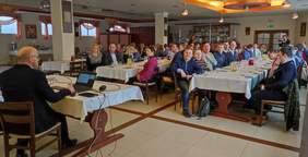 Pokaż zdjęcie: Na dużej Sali bankietowej przy stole na którym stoi laptop siedzi mężczyzna w dłoni trzyma mikrofon przed nim przy dwóch długich stołach siedzą słuchacze spotkania.