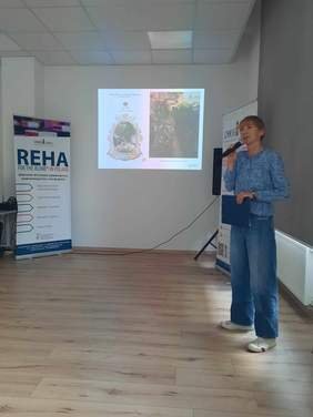 Pokaż zdjęcie: Kobieta ubrana na niebiesko stoi i przemawia do mikrofonu. Na ścianie za nią wyświetlona jest prezentacja. Po bokach znajdują się banery Fundacji Szansa. 