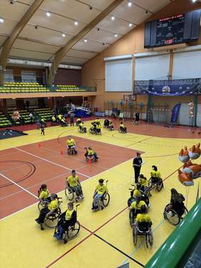 Pokaż zdjęcie: Widok hali sportowej z góry. Na boisku znajduje się grupa osób poruszających się na wózkach, ustawionych w kilku rzędach pomiędzy pachołkami. W tle widoczne są trybuny oraz tablica wyników