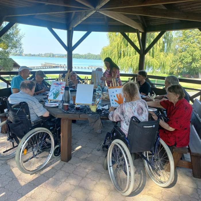 Pokaż zdjęcie: Grupa osób z niepełnosprawnościami, wśród nich osoby na wózku, znajduje się w domku letniskowym i odbywa zajęcia rehabilitacyjne