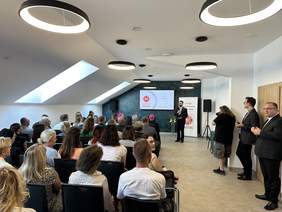 Pokaż zdjęcie: Na Sali stoi dużo krzeseł w kilku rzędach na których siedzą na galowo ubrani goście. Przed nimi stoi ksiądz trzymający mikrofon.