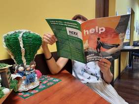 Pokaż zdjęcie: W korytarzu siedzi na krześle kobieta, która trzyma w dłoniach miesięcznik „Help – jesteśmy razem”. Jedną rękę ma opartą na stole, na którym leżą różne kolorowe dekoracje. 