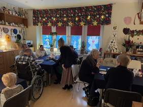 Pokaż zdjęcie: Grupa osób spędza czas w przytulnym, świątecznie udekorowanym pomieszczeniu. Przy stole siedzą goście, w tym mężczyzna na wózku. W tle choinka, stroiki i kolorowe dekoracje na oknach tworzą ciepły, świąteczny klimat.