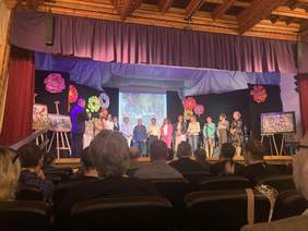 Pokaż zdjęcie: Na scenie stoi kilkunastu uczestników jubileuszu, w tle na ekranie kadr z filmu przedstawiającego w skrócie 25 lat działalności placówki