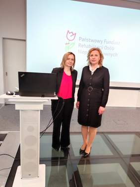 Pokaż zdjęcie: Przy mównicy stoją dwie kobiety, od lewej kobieta blondynka w ciemnym garniturze, po prawej kobieta blondynka w czarnej sukience.