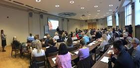 Pokaż zdjęcie: Grupa kilkudziesięciu osób siedzi w sali konferencyjnej i przysłuchuje się wystąpieniu jednego z prelegentów. Obok dużego ekranu, na którym wyświetlona jest prezentacja, ustawiony jest baner PFRON, a po lewej stronie stoi kobieta z telefonem w dłoni.