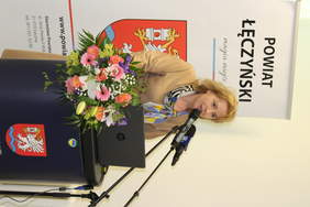 Pokaż zdjęcie: Przy mównicy stoi kobieta w jasnej beżowej sukience, na mównicy stoi laptop oraz leży bukiet kolorowych kwiatów