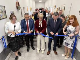 Pokaż zdjęcie: Uroczyste przecięcie niebieskiej wstęgi przez pięć osób w eleganckich strojach, otoczone grupą uczestników wydarzenia w jasnym, udekorowanym korytarzu z obrazami na ścianach.