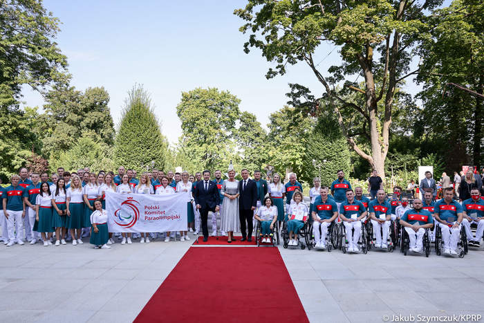 Pokaż zdjęcie: kilkadziesiąt osób stoi na tle parku pozując do fotografii, wszyscy w strojach sportowych. Na środku stoi para prezydencka w eleganckich strojach. Prowadzi do niej czerwony dywan.