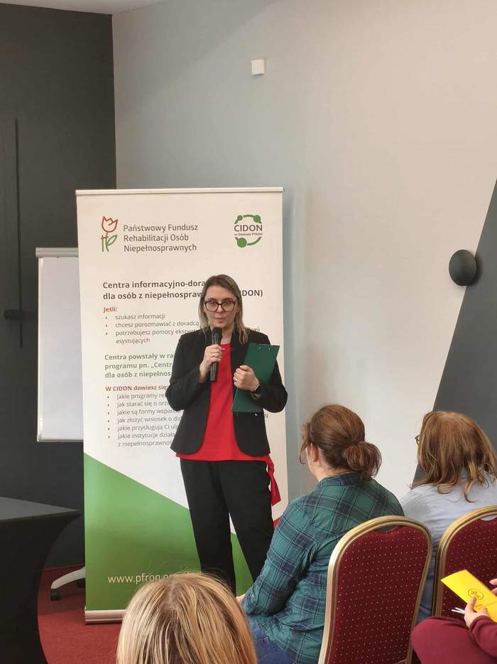 Kobieta w czarnym garniturze i czerwonej bluzce, stoi, za nią biało-zielono-czerwony baner