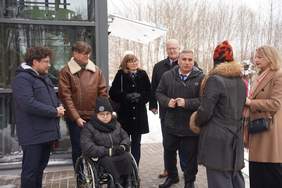 Pokaż zdjęcie: Grupa osób stojących przed przeszkloną windą zewnętrzną przy budynku. Wśród uczestników osoba poruszająca się na wózku inwalidzkim, wszyscy ubrani w zimowe płaszcze, wokół śnieg i brukowany chodnik.