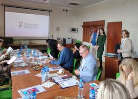 Pokaż zdjęcie: Po prawej i lewej stronie długiego stołu siedzą uczestnicy spotkania. Przed nimi na stole leżą broszury. Na ekranie wyświetla się slajd z napisem i logo PFRON. Po prawej stronie sali stoją dwie kobiety.