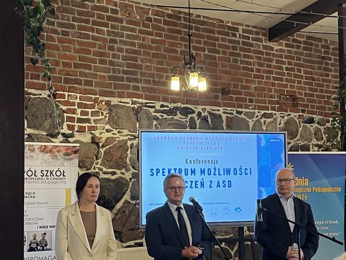 Na środku sali stoją trzy osoby - od lewej kobieta i dwóch mężczyzn. Za nimi widać ekran, z wyświetlonym tytułem konferencji