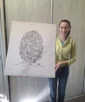 Pokaż zdjęcie: Na szarym, prostokątnym płótnie wyhaftowano czarną nicią drzewo o gęstej, okrągłej koronie. Jej wnętrze wypełniają drobne motywy roślinne – liczne liście i kwiaty przypominające róże – oraz małe ptaki siedzące na gałązkach. Smukły pień wyrasta z dwóch łagodnych pagórków zaznaczonych pojedynczą linią; na lewym widać trzy małe krzaczki, a na prawym dwa. Tło pozostaje jednolite i surowe. Obok obrazu stoi jego autorka – uśmiechnięta kobieta o jasnej cerze i ciemnych, upiętych włosach. Ma na sobie jasnozielony sweter oraz niebieskie dżinsy.
