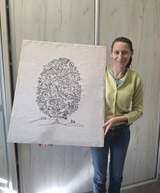 Na szarym, prostokątnym płótnie wyhaftowano czarną nicią drzewo o gęstej, okrągłej koronie. Jej wnętrze wypełniają drobne motywy roślinne – liczne liście i kwiaty przypominające róże – oraz małe ptaki siedzące na gałązkach. Smukły pień wyrasta z dwóch łagodnych pagórków zaznaczonych pojedynczą linią; na lewym widać trzy małe krzaczki, a na prawym dwa. Tło pozostaje jednolite i surowe. Obok obrazu stoi jego autorka – uśmiechnięta kobieta o jasnej cerze i ciemnych, upiętych włosach. Ma na sobie jasnozielony sweter oraz niebieskie dżinsy.