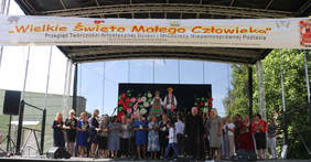 Pokaż zdjęcie: Na scenie stoi grupa kobiet nad nimi wisi napis Wielkie Święto Małego Człowieka” Przeglądem Twórczości Artystycznej Dzieci i Młodzieży Niepełnosprawnej Podlasia.