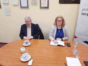 Pokaż zdjęcie: Przy stole siedzi dyrektor Łódzkiego Oddziału PFRON oraz ekspertka z CIDON. Kobieta notuje coś w terminarzu, a mężczyzn rozmawia z interlokutorami. Na stole leżą filiżanki, tefefony, notatki i długopisy. 