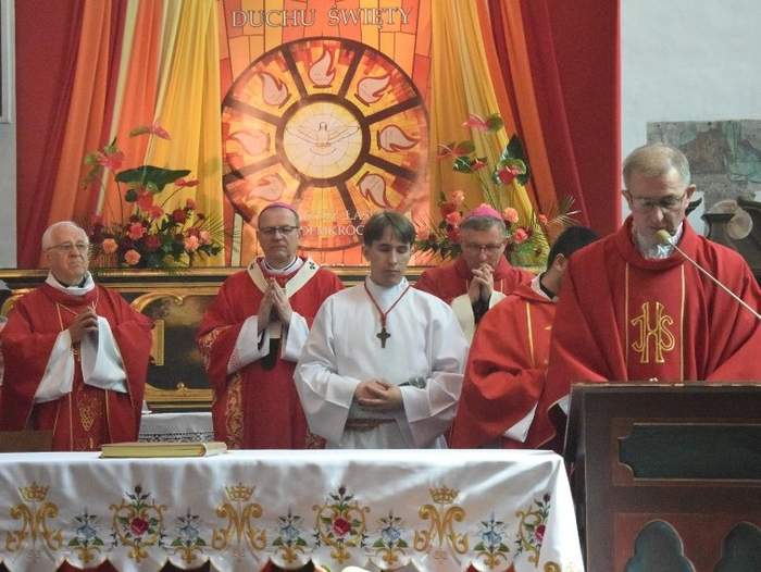 Pokaż zdjęcie: Za ołtarzem stoi kilku księży w uroczystych czerwonych szatach. Przed mównicą ksiądz w okularach kruczych włosach mówi do jasnego mikrofonu. W tle symbol i napis Duch Święty.