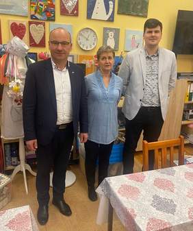 Pokaż zdjęcie: Kobieta i dwóch mężczyzn w garniturach znajdują się w kolorowym pomieszczeniu, za nimi ściana z obrazkami i regał z artykułami plastycznymi