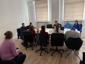 Pokaż zdjęcie: W jasnym pomieszczeniu biurowym grupa osób siedzi wokół owalnego stołu, prawdopodobnie biorąc udział w spotkaniu lub dyskusji. Większość osób jest zwrócona w stronę stołu.